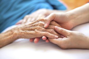 An elderly woman holding the hand of a nurse while she lays in bed at at cancer treatment center.