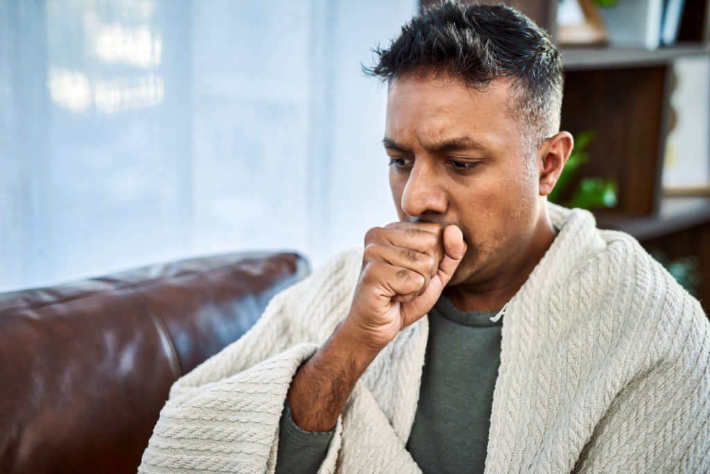 A man wrapped up in a sweater sitting down on his couch coughing due to his upper respiratory infection.