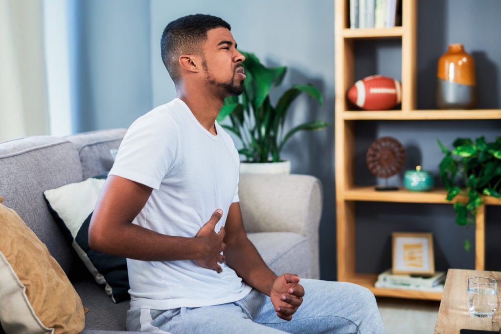 A man sitting on the couch holding his stomach due to abdominal pain.