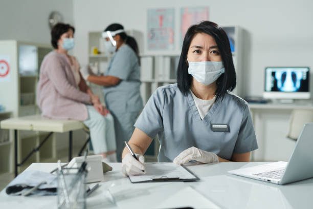 Young brunette female clinician in protective workwear sitting by desk in front of camera while her colleague vaccinating patient on background