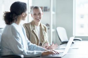 Two women smile as they discuss with one another. One of the young women has a shaved head in cancer center